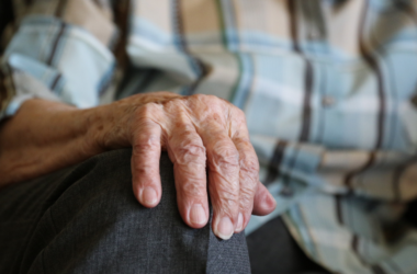 Mano de una persona mayor apoyada sobre su rodilla, con camisa de cuadros y pantalón gris