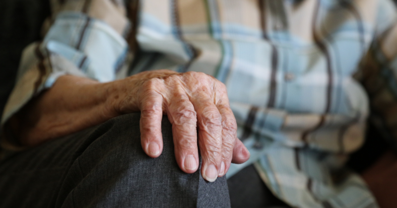 Mano de una persona mayor apoyada sobre su rodilla, con camisa de cuadros y pantalón gris