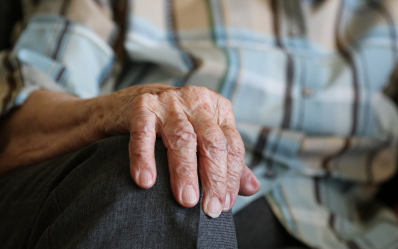 Mano de una persona mayor apoyada sobre su rodilla, con camisa de cuadros y pantalón gris