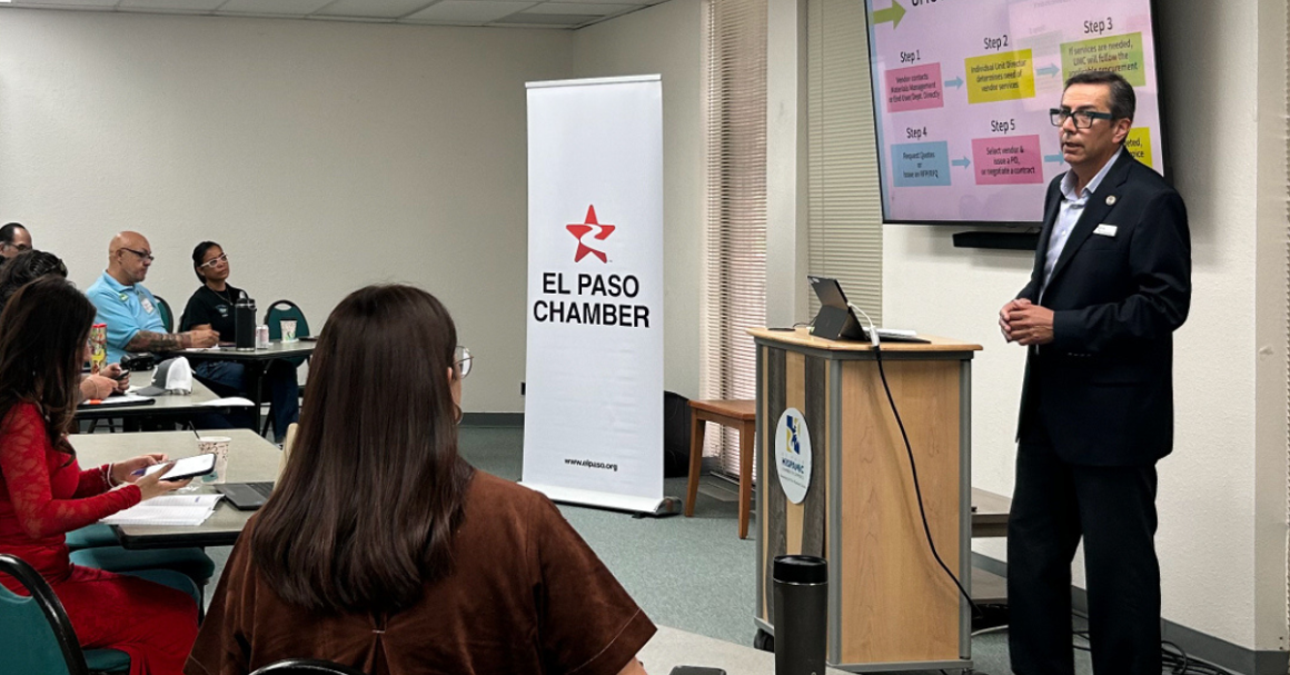 Hombre presenta ante un grupo de asistentes junto a un banner de El Paso Chamber y una pantalla con un diagrama por pasos