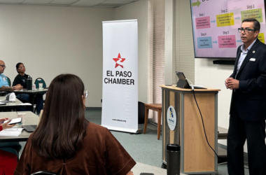 Hombre presenta ante un grupo de asistentes junto a un banner de El Paso Chamber y una pantalla con un diagrama por pasos