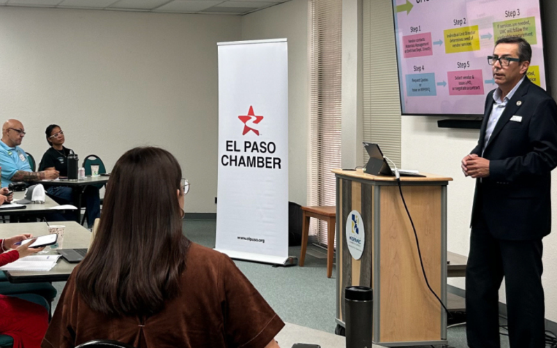 Hombre presenta ante un grupo de asistentes junto a un banner de El Paso Chamber y una pantalla con un diagrama por pasos